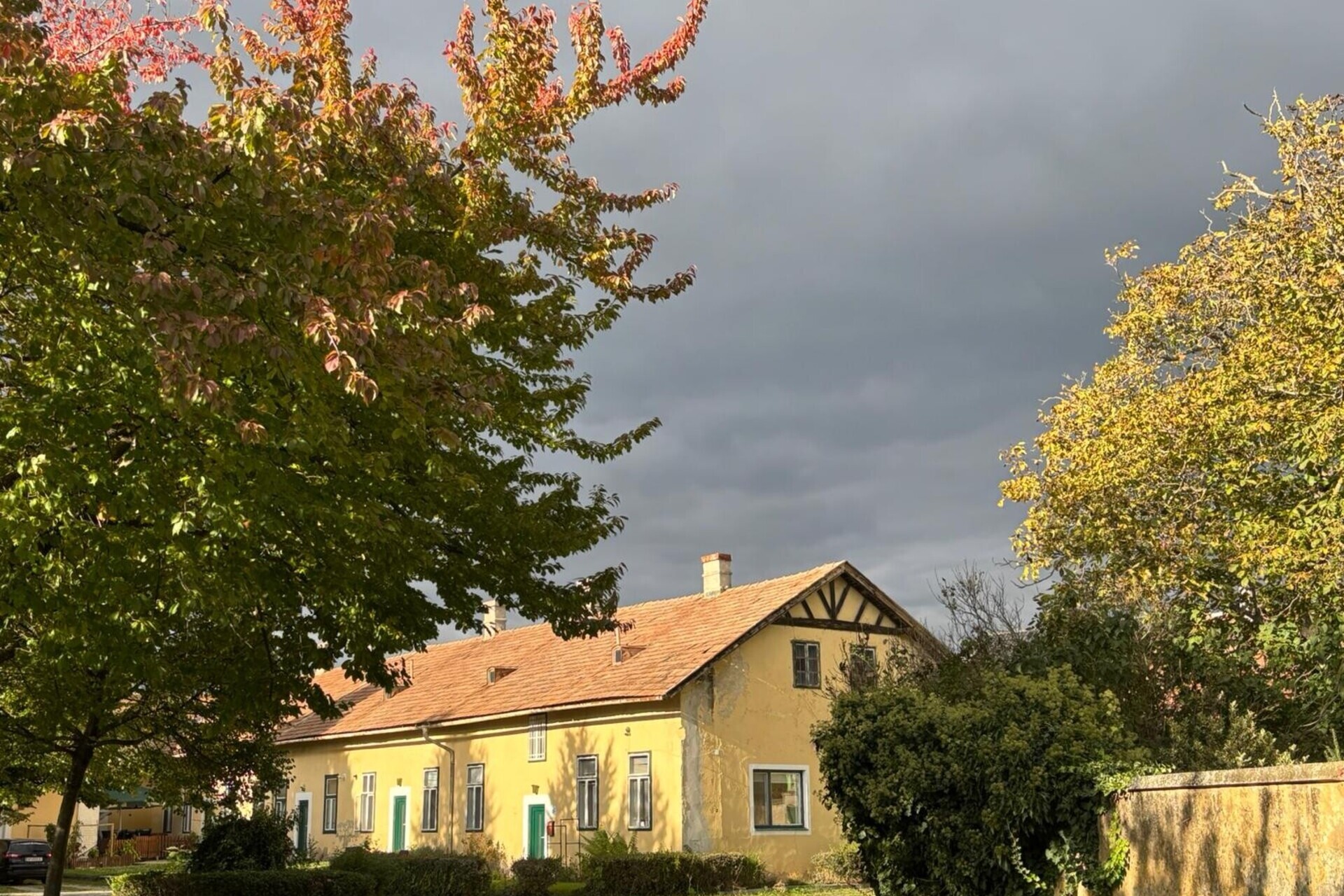 Hier siehst du das Gebäude von Yoga am Hof von aussen in toller Herbststimmung mit dunklem Himmel und Bäumen mit bunten Blättern im Vordergrund.