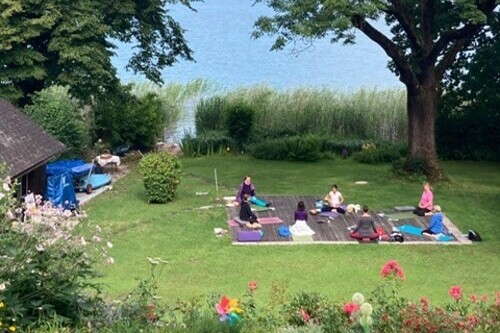 Garten voller Blumen und grün mit See und toller Stimmung. Auf einer Holzplattform ist eine bunte Yogagruppe versammelt.